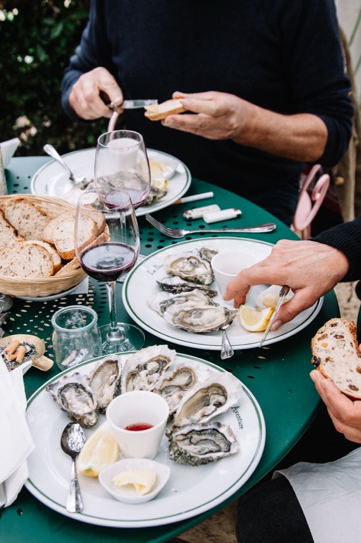 Deux assiettes d’huîtres, une corbeille de pain et du vin rouge sur la table verte, mains en train de servir