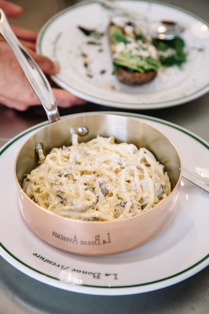 Des pâtes servies dans une casserole en cuivre gravée au nom du lieu, posée sur une assiette siglée