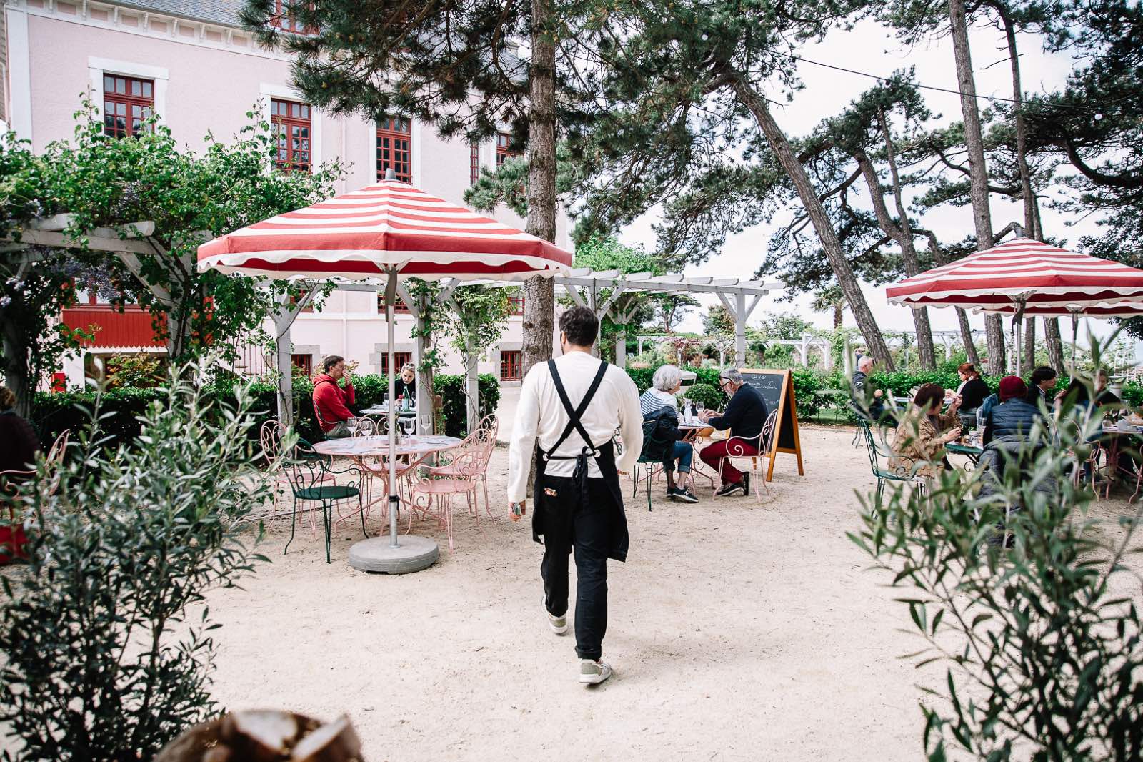 La terrasse du salon de thé : parasols rayés rouge et blanc, chaises roses en fer et, au centre de l’allée, un serveur qui s’éloigne