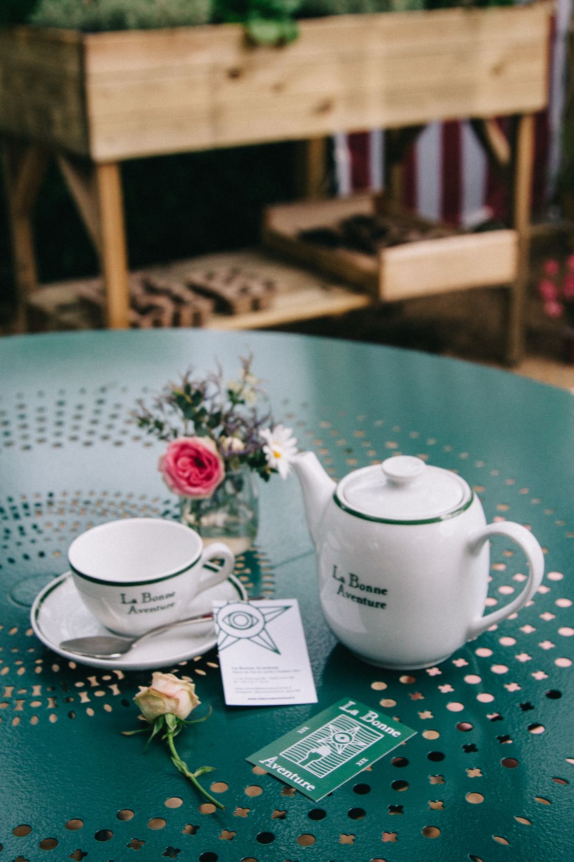 Théière et tasse siglées La Bonne Aventure sur une table de jardin vert foncé, avec les deux cartes et une rose