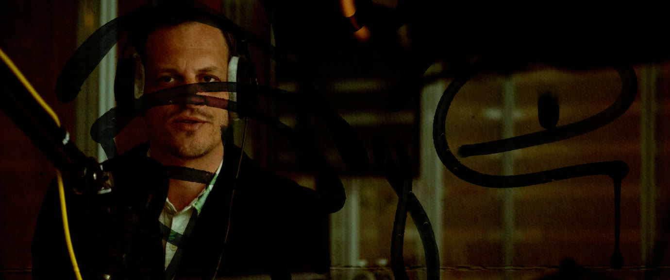 En studio d’enregistrement, casque sur les oreilles, derrière une vitre taguée
